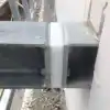 Witte kunststofplaat tussen twee grijze metalen kokerprofielen, vastgezet met bouten, tegen een witte muur op een bouwplaats.