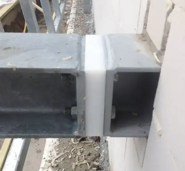 Witte kunststofplaat tussen twee grijze metalen kokerprofielen, vastgezet met bouten, tegen een witte muur op een bouwplaats.