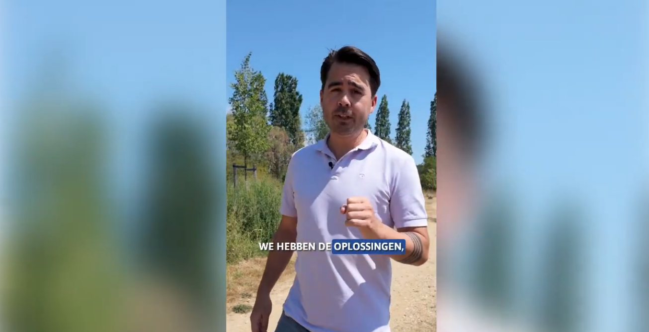Man in een lichtpaars poloshirt spreekt buiten op een zandpad, met bomen en een blauwe lucht op de achtergrond.